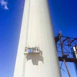 Asbestos removal on a tall cycliner stack. Construction rafting is rigged in place with mutple workers standing on it. Bright blue sky in the background and a stell structure on the right side.